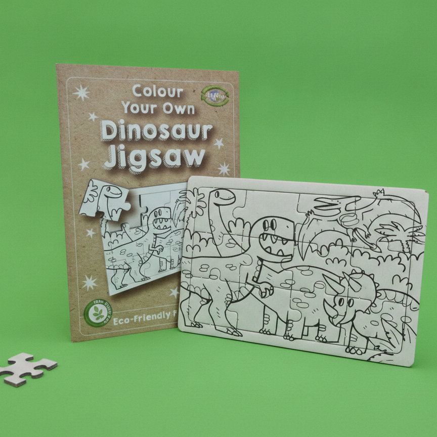 A children's dinosaur jigsaw puzzle set on a bright green background, showing the brown cardboard cover, the assembled black-and-white puzzle sheet, and one loose puzzle piece.