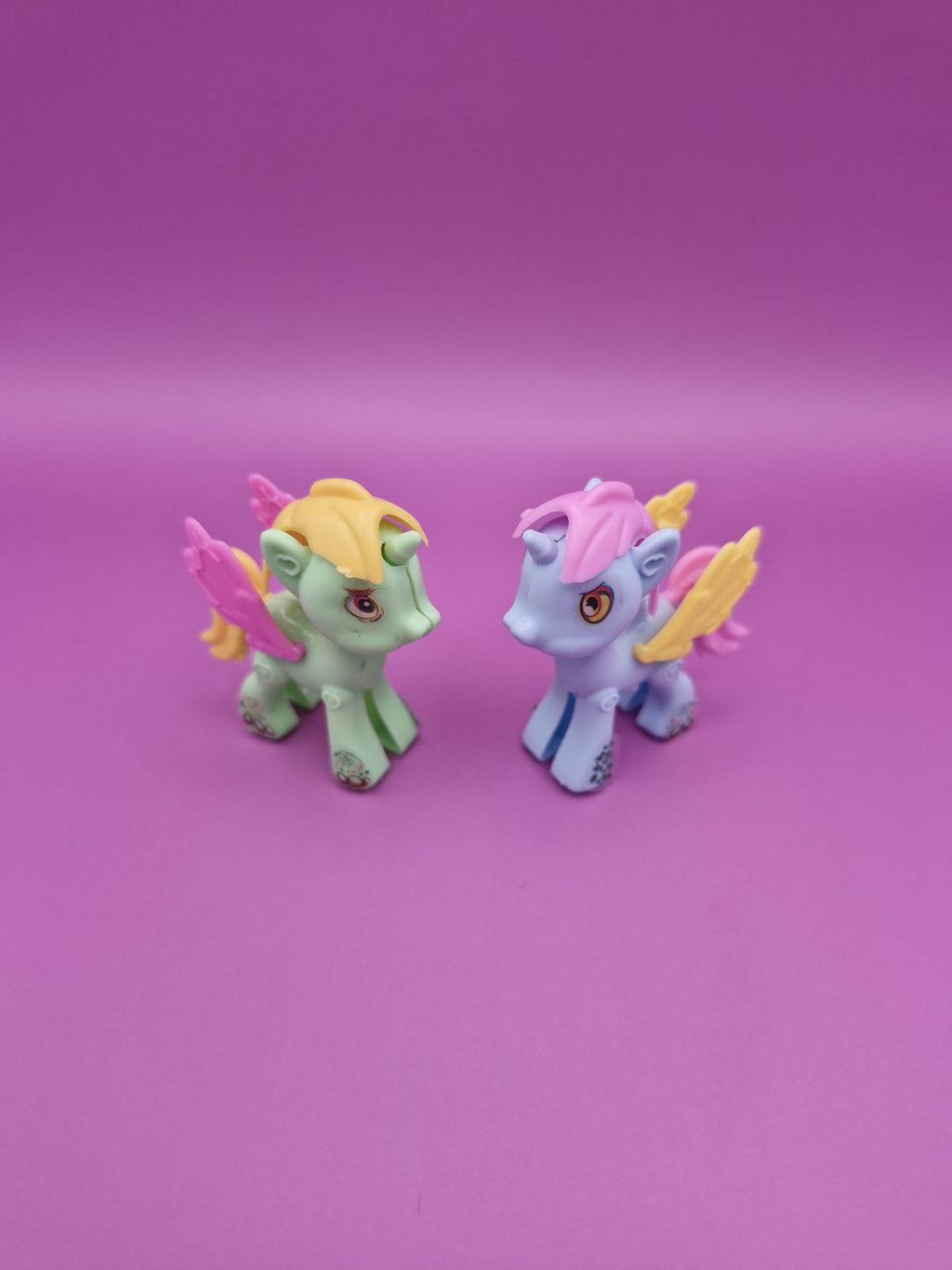 Two toy unicorns with colourful hair and wings on a pink background