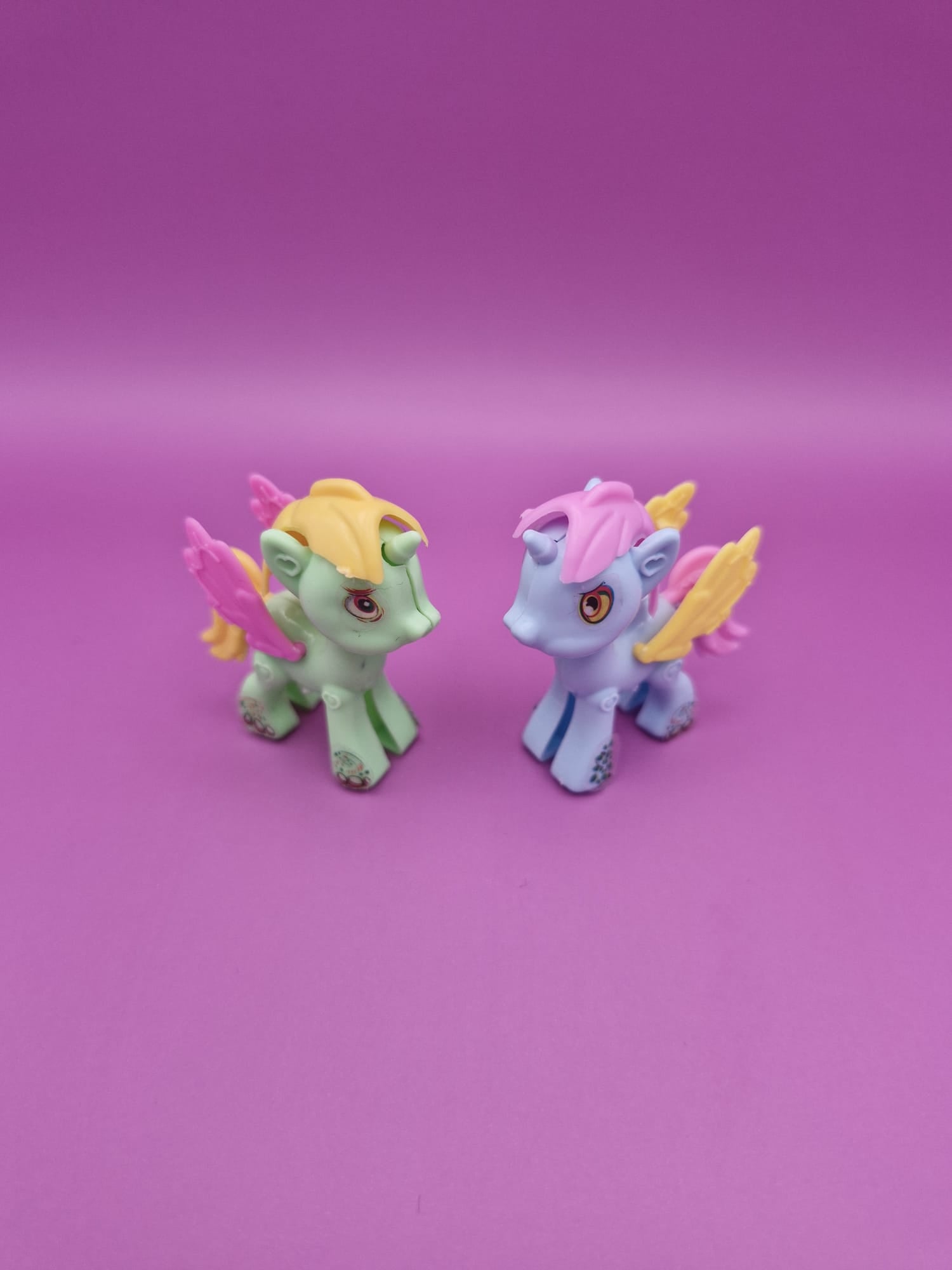 Two toy unicorns with colourful hair and wings on a pink background