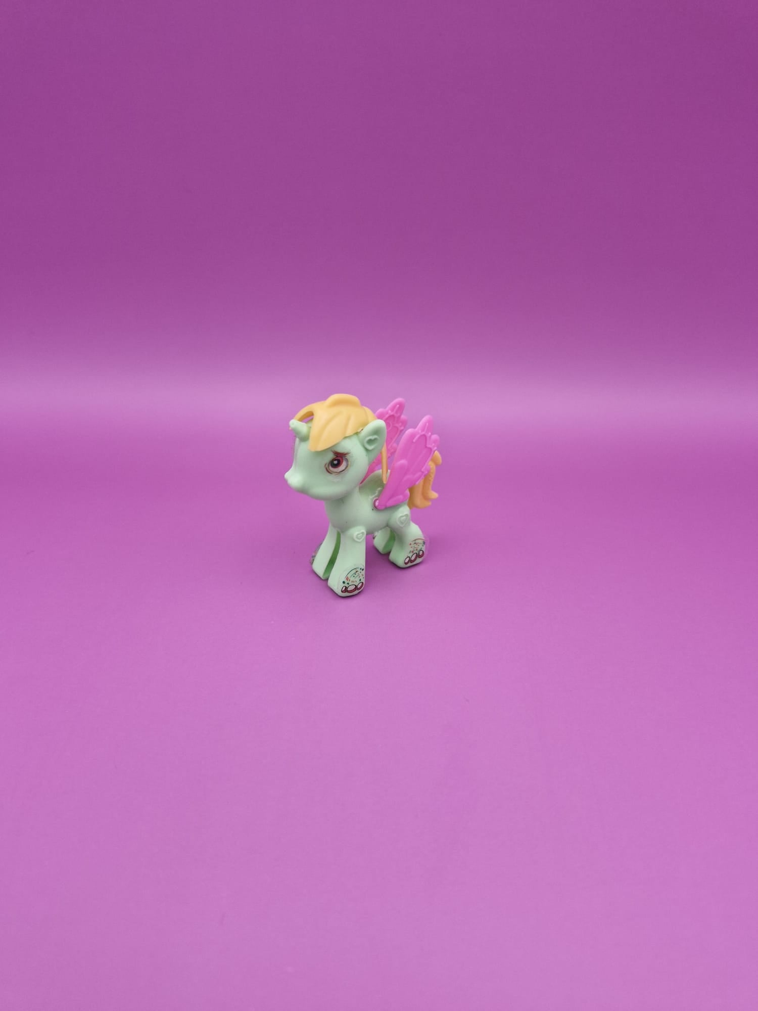 Small green unicorn figurine with yellow hair and pink wings on a purple background.